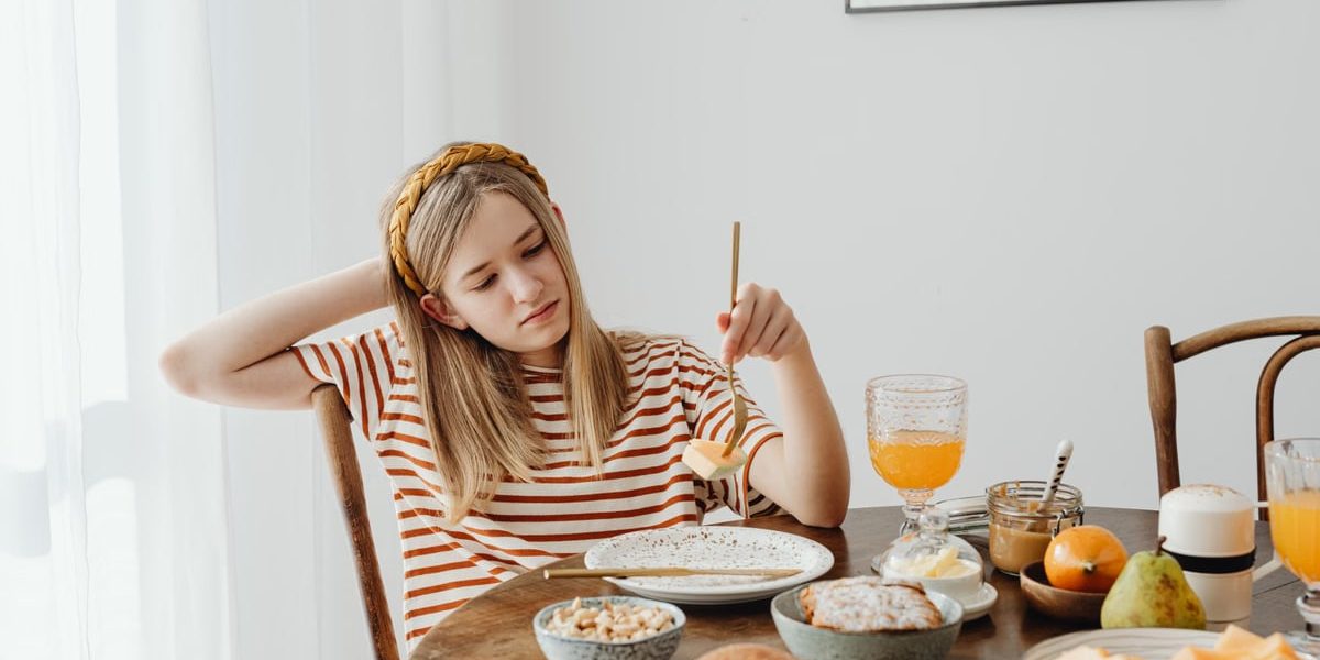 Teen girl in striped shirt at breakfast table with fruits, bread, and juice. Casual morning setting.